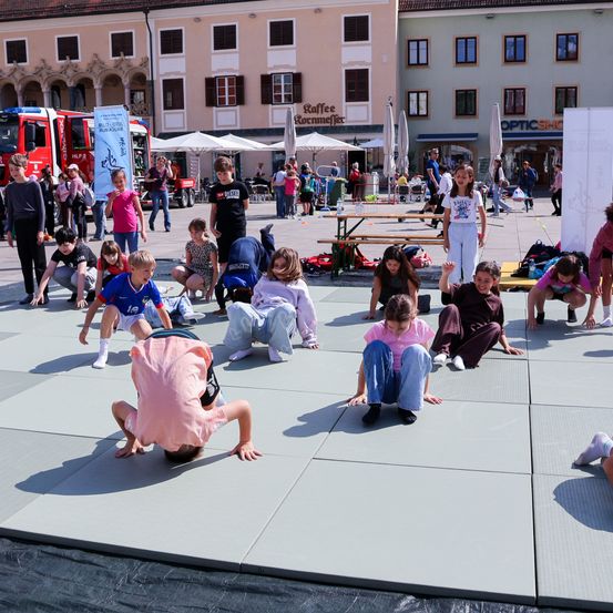 Eine Gruppe von Kindern macht Yoga-Posen auf einem gepflasterten Bereich in einer Stadtmitte. In der Nähe sind Gebäude, ein roter Feuerwehrwagen und Tische mit Stühlen.