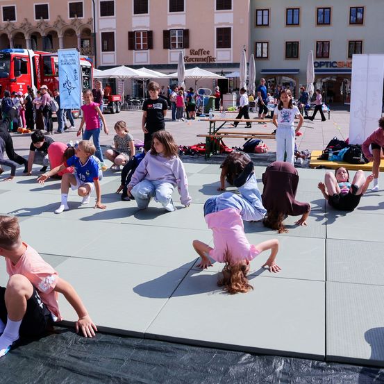 Mehrere Kinder machen Yoga-Posen auf einem Stadtplatz. Im Hintergrund ist ein roter Feuerwehrwagen und ein Gebäude mit dem Schild 'Raffee' zu sehen. Viele Menschen gehen in der Gegend umher.