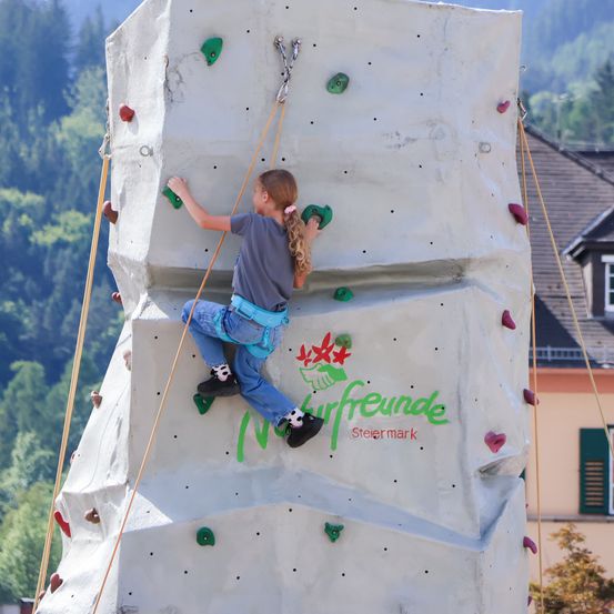 Ein junges Mädchen in einem grauen T-Shirt und Jeans klettert an einer weißen Kletterwand mit grünen und roten Griffen. Die Wand hat das Wort "Stiermark" in Grün. Hinter ihr ist ein Haus mit braunem Dach und grünen Fensterläden zu sehen.