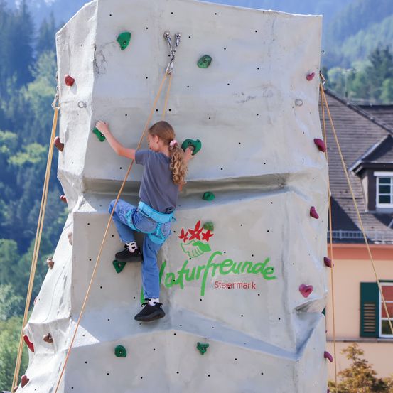 Ein kleines Mädchen klettert an einer Kletterwand mit einem Haus und Bäumen im Hintergrund.
