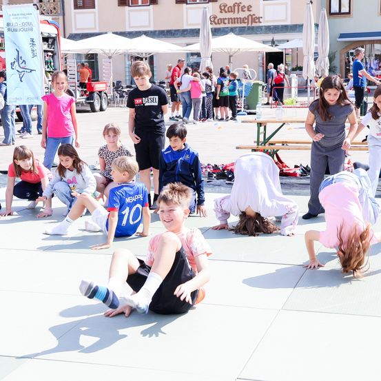 Kinder führen an einem sonnigen Tag gymnastische Übungen durch und tragen verschiedene Sporttrikots. Einige Erwachsene stehen und beobachten. Im Hintergrund befinden sich Zelte, Schirme und ein Gebäude mit einem Schild.