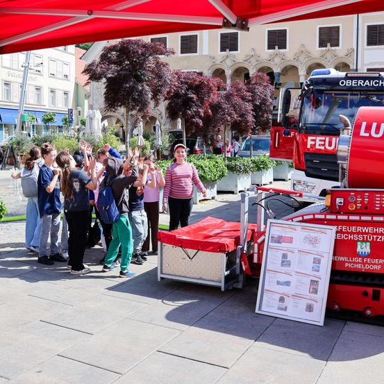 Eine Gruppe von Kindern winkt einem roten Feuerwehrwagen mit einem roten Vordach und Schild vor einem Gebäude zu.