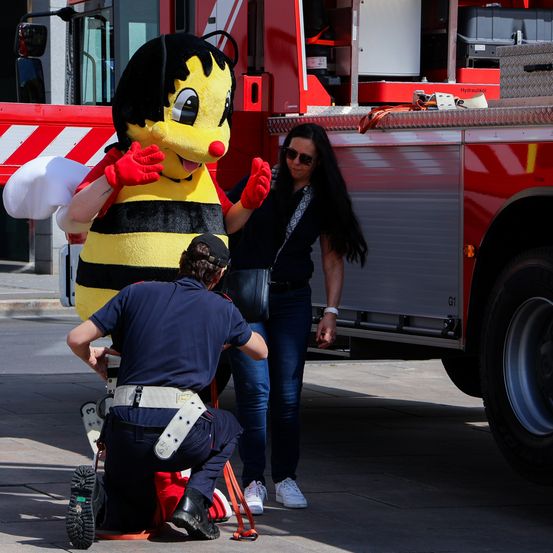 Ein Mann in Uniform kniet vor einem Feuerwehrauto, eine Frau steht hinter einer Maskottchen.