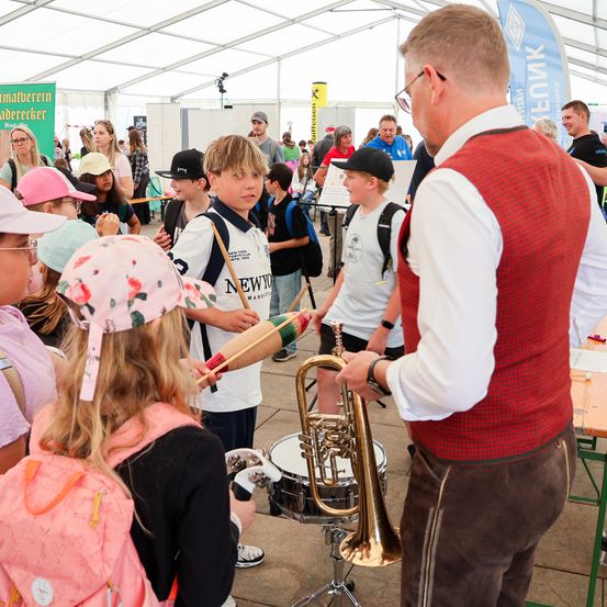 Ein Mann zeigt Kindern, wie man Trommeln und Trompete spielt, bei einer Outdoor-Veranstaltung. Hinter ihnen steht eine Menschenmenge und schaut zu. Im Hintergrund sind Banner zu sehen.