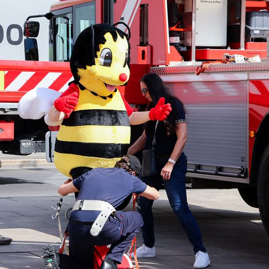 Ein Bienenmaskottchen interagiert mit einer Frau und einem Feuerwehrmann an einem sonnigen Tag. Der Feuerwehrmann kniet, die Frau steht und sie alle schauen auf die Biene. Hinter ihnen steht ein roter Feuerwehrwagen.