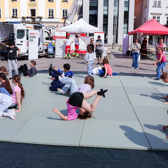 Eine Gruppe von Kindern spielt auf einer erhöhten Matte in einem Außenbereich, während einige Erwachsene zusehen. Ein weißer Lieferwagen mit den Worten 'WIR SIND DA' ist in der Nähe geparkt.
