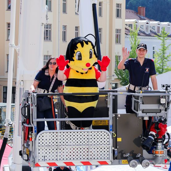 Ein Feuerwehrmann in Uniform und eine Frau mit Brille winken von einem Feuerwehrwagen, mit einem großen Bienen-Maskottchen in der Mitte.