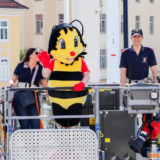 Ein Maskottchen in einem Bienenkostüm winkt auf einer Plattform mit einer Frau und einem Mann in Uniform. Dahinter steht ein Gebäude mit vielen Fenstern.