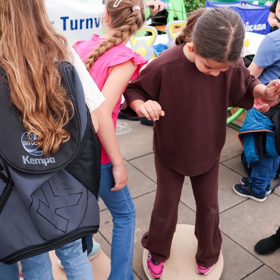 Ein Mädchen in brauner Hose steht auf einem Balancebrett und schaut nach unten. Ein Mädchen mit Rucksack steht hinter ihr, und eine weitere Mädchen ist links von ihr. Im Hintergrund stehen und sitzen Menschen um einen Tisch.