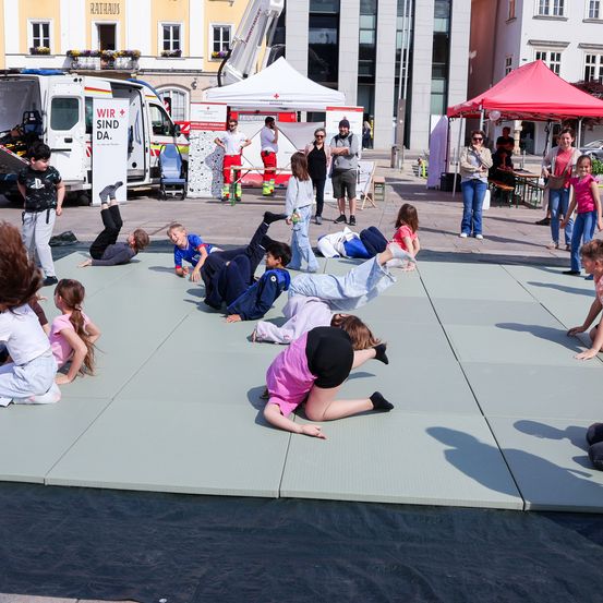 Kinder machen Gymnastik auf Matten in einem offenen Bereich. Ein Krankenwagen und Zelte im Hintergrund. Menschen stehen und beobachten.