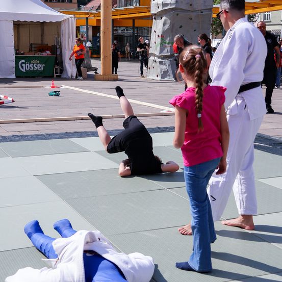 Ein Kampfkunstunterricht findet statt, bei dem eine Person einen Handstand macht. In der Nähe beobachtet ein Mädchen, während ein Mann anweist. Die Szene spielt sich auf einem gefliesten Boden ab, mit einem Zelt und einer Kletterwand im Hintergrund.
