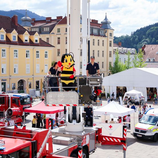 Ein Bienen-Maskottchen steht auf einer Feuerwehrplattform mit zwei Personen, umgeben von Zelten und Gebäuden, mit Rettungsfahrzeugen und Menschen im Hintergrund.