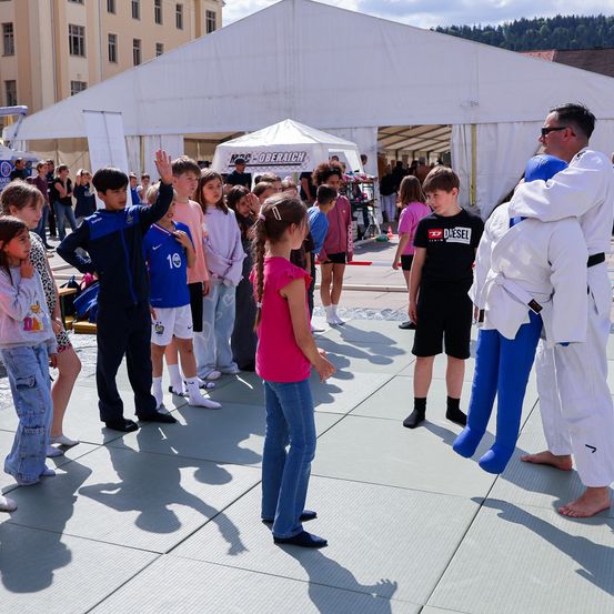 Eine Gruppe von Menschen, darunter Kinder, stehen in einer Schlange auf einer Bühne. Ein Mann unterrichtet einen Jungen mit einem Dummy in Judo. Hinter ihnen schauen viele Leute zu.