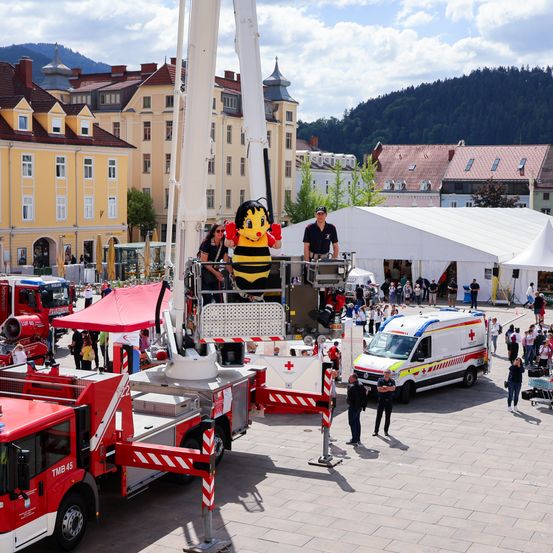 Auf einem Stadtplatz steht ein Feuerwehrauto mit einer Biene als Maskottchen auf seiner Plattform, umgeben von Menschen. Ein rotes Zelt befindet sich in der Nähe. Andere Fahrzeuge, einschließlich eines Krankenwagens, sind ebenfalls anwesend. Im Hintergrund sind Gebäude mit roten Dächern zu sehen.