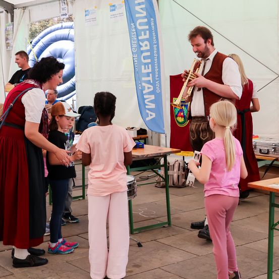 Ein Mann spielt Trompete, während zwei Mädchen und ein Junge zuschauen. Eine erwachsene Frau steht hinter ihnen und spricht mit den Kindern. Ein Banner steht 'Annalenbeima'.
