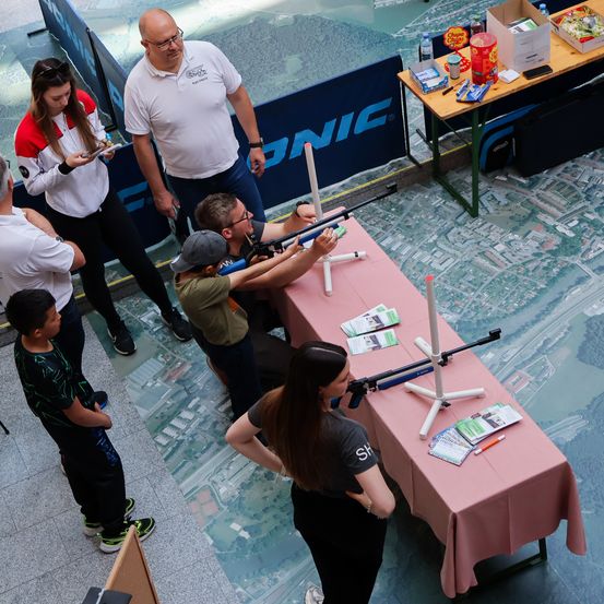 Ein Indoor-Schießstand mit Teilnehmern, zwei vor einem pinkfarbenen Tisch mit einem Gewehr, andere beobachten, und einem blauen Hintergrund mit Text.