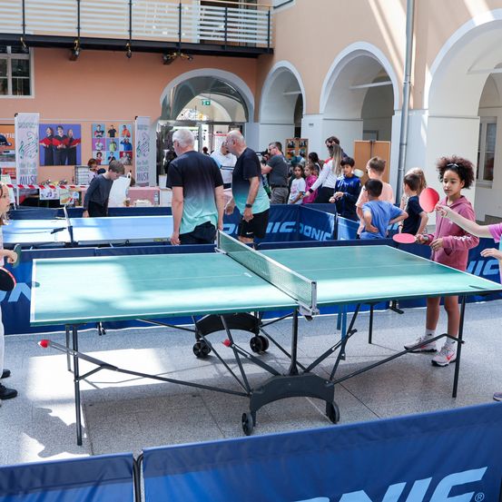 Eine Gruppe von Menschen spielt drinnen Tischtennis mit zwei aufgestellten Tischen. Zuschauer beobachten, und einige Banner sind sichtbar.