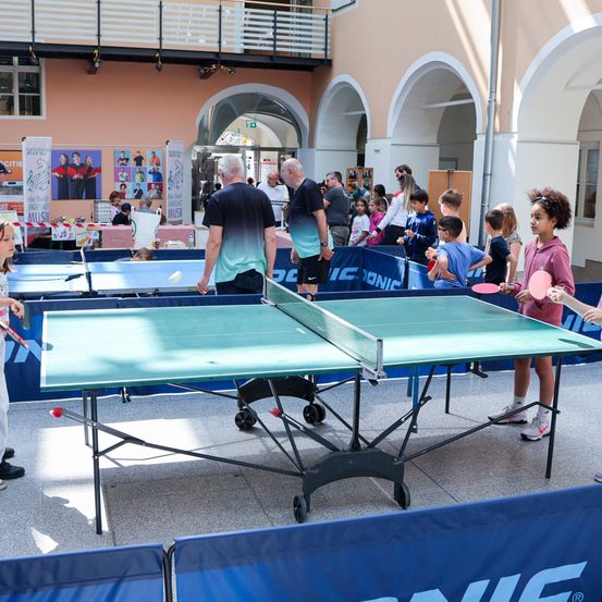 Eine Gruppe von Menschen spielt Tischtennis in Innenräumen. Zwei Kinder schlagen den Ball hin und her. Zuschauer beobachten von den Seiten. Banner sind an der Wand, und eine Terrasse ist darüber.
