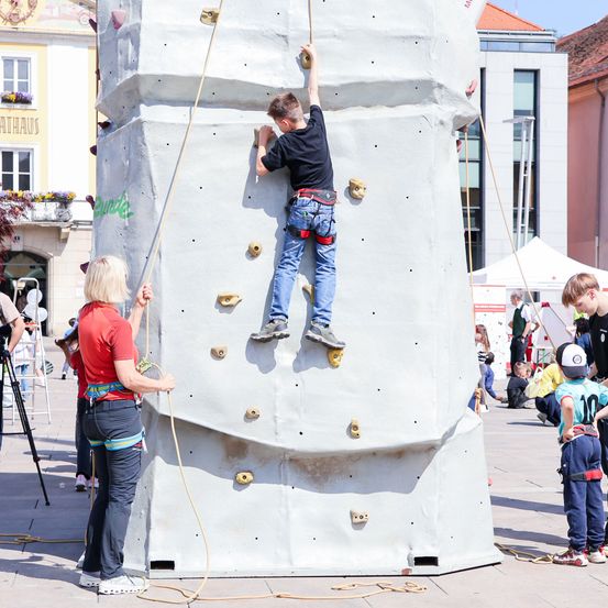 Ein junger Junge klettert an einer künstlichen Kletterwand, während eine Frau hilft. Mehrere andere beobachten. Im Hintergrund sind Gebäude und Zelte zu sehen.