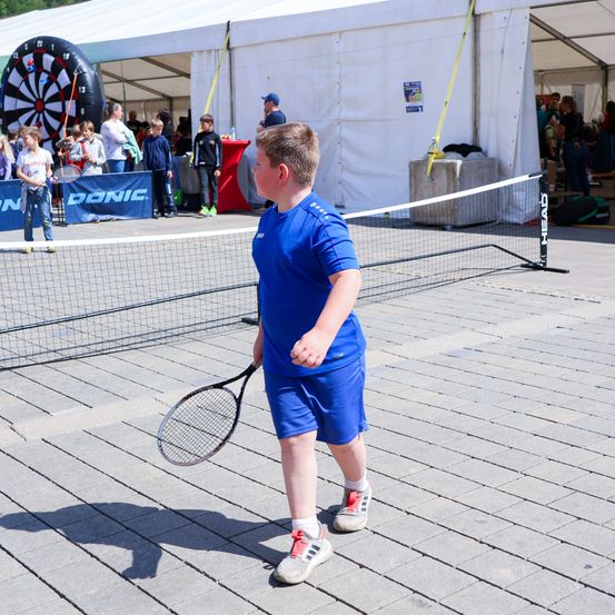 Ein junger Junge hält eine Tennisschläger auf einem Tennisplatz. Mehrere Menschen stehen hinter ihm, und ein großes Dartbrett ist sichtbar.
