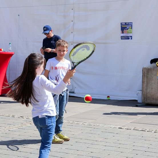 Ein junges Mädchen schwingt einen Tennisschläger, gefolgt von einem Jungen. Ein Mann beobachtet im Hintergrund. Tennisbälle sind auf dem Boden verstreut.