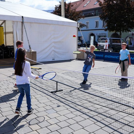 Vier Kinder spielen Tennis auf einem kleinen Platz mit Netz in einem Außenbereich, zwei von ihnen schlagen den Ball über das Netz, während die anderen zusehen. Ein Zelt und einige Gebäude sind im Hintergrund.