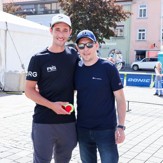 Zwei Männer stehen auf einem Stadtplatz. Einer trägt eine Kappe und hält einen Tennisball. Der andere trägt eine Sonnenbrille und eine Uhr. Dahinter befindet sich ein Zelt, ein Gebäude und ein geparktes Auto.