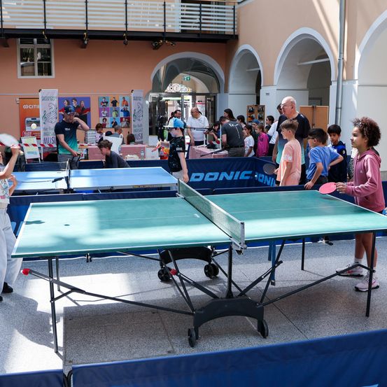 Menschen spielen Tischtennis in einer Halle. Einige schauen zu. Die Tische haben blaue Hintergründe und ein Markenlogo. Eine Frau hält eine Schläger.