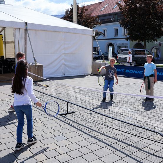 Eine Gruppe von Kindern spielt Tennis auf einem Ziegelplatz. Der Platz hat ein Netz und ein Zelt im Hintergrund. Ein Kind ist dabei, den Ball zu schlagen.