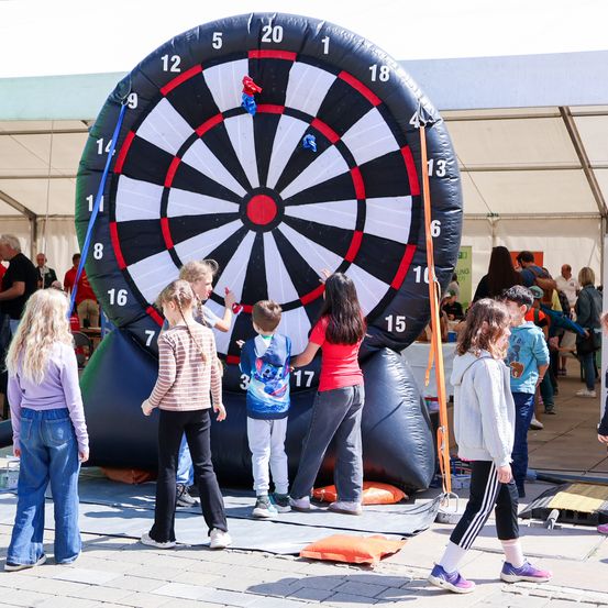 Mehrere Kinder und Erwachsene spielen ein riesiges Dartspiel bei einer Outdoor-Veranstaltung. Das Dartbrett ist aufgeblasen und farbenfroh.