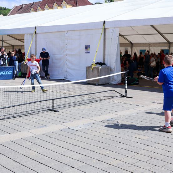 Zwei Kinder spielen Tennis auf einem Platz unter einem weißen Zelt. Einer hält eine Schläger, der andere schwingt sie. Zuschauer beobachten in der Nähe, einige stehend und andere sitzend.