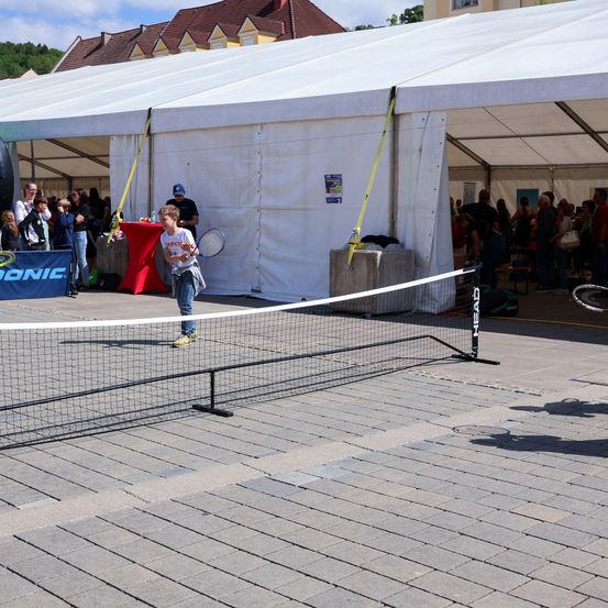 Ein Junge spielt Tennis mit einer Menge zuschauender Menschen unter einem Zelt. Der Tennisplatz ist von einem Netz umgeben.