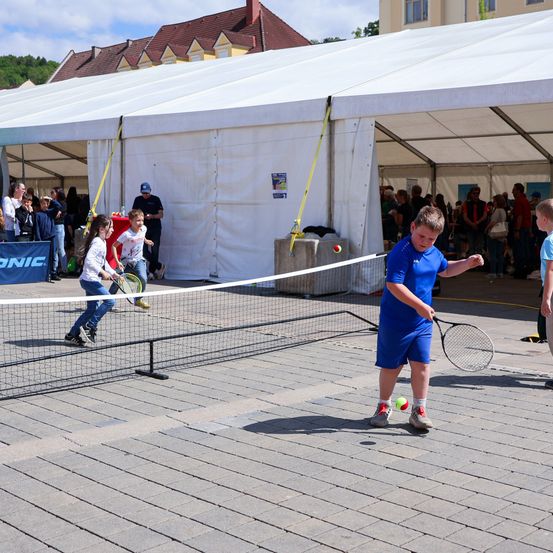 Eine Gruppe von Kindern spielt Tennis unter einem Zelt im öffentlichen Raum. Ein Junge in Blau bereitet sich darauf vor, den Ball zu schlagen.