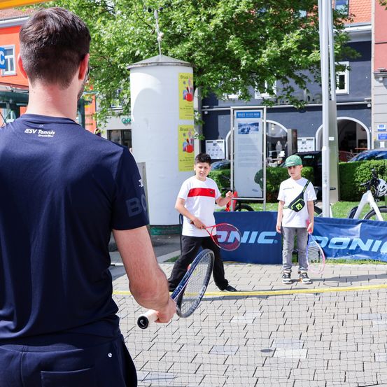 Ein Mann beobachtet zwei Jungen, die auf einem Tennisplatz spielen. Die Jungen halten Schläger und einer trägt einen Hut. Im Hintergrund sind ein blaues Netz und ein blauer Banner sichtbar.