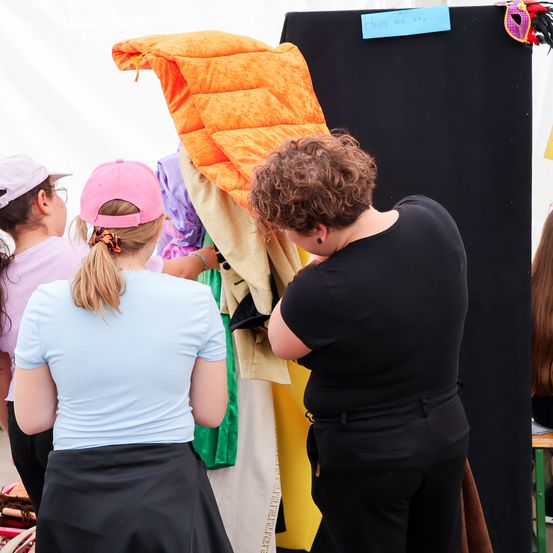 Drei Frauen und ein Kind versammeln sich um einen Kleiderständer mit Kleidung und Kostümen, eine Person justiert die orange Quiltdecke.