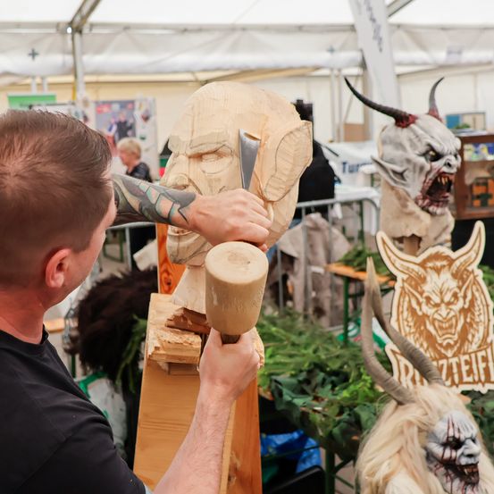 Ein Mann schnitzt ein Gesicht aus Holz mit einem Hammer und Meißel. Er hat Tätowierungen am Arm. Im Hintergrund sind weitere Holzskulpturen ausgestellt.