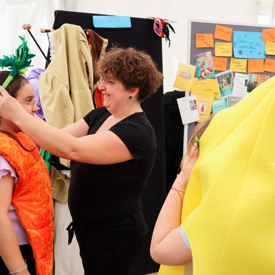 Eine Frau passt das Kostüm eines Kindes an, das eine orange Weste und ein Bananenkostüm trägt. Dahinter ist eine Pinnwand mit bunten Papieren bedeckt.