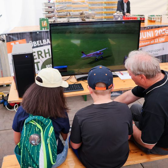 Drei Personen sehen sich ein Flugsimulator auf einem Monitor an. Ein Mann und zwei Kinder, eines mit Rucksack, sitzen auf einer Bank. Der Bildschirm zeigt ein Flugzeug.