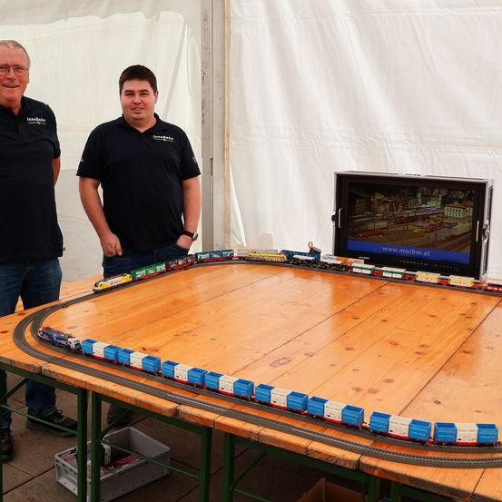 Zwei Männer stehen neben einem großen Holztisch mit einer Modelleisenbahn. Der Bildschirm hinter ihnen zeigt ein Video einer Stadt.
