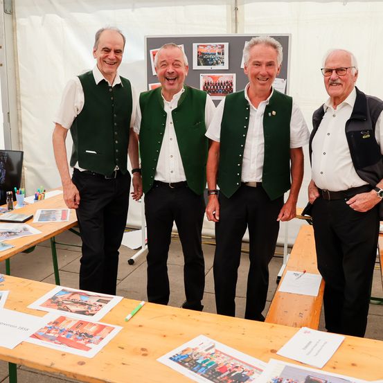 Vier ältere Männer lächeln und posieren für ein Foto. Sie stehen in einer Reihe vor einem Tisch mit Papieren und Stiften. Dahinter befindet sich eine Tafel mit Bildern. Sie tragen alle grüne Westen und weiße Hemden.