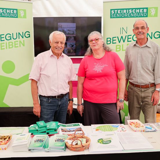 Drei Personen stehen hinter einem Tisch. Sie lächeln und scheinen auf einer Messe zu sein. Auf dem Tisch liegen Broschüren und Flyer. Hinter ihnen steht ein grüner Banner mit weißem Text.