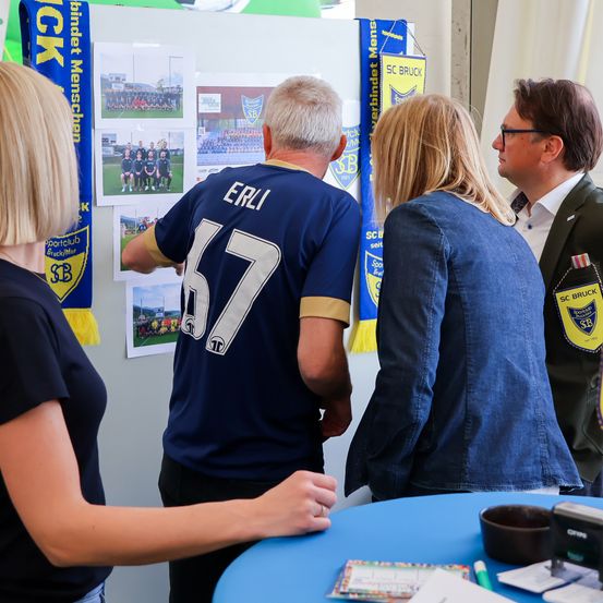 Vier Menschen stehen um einen Tisch herum, zwei Frauen und zwei Männer, in Trikots von Sportvereinen. Der Mann auf der linken Seite trägt ein blaues Shirt mit der Nummer 57. An der Wand dahinter hängen Fotos von Fußballmannschaften und Banner.