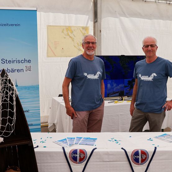 Zwei Männer stehen hinter einem Tisch mit Broschüren bei einer Outdoor-Veranstaltung. Ein Mann hat einen Bart, der andere trägt eine Brille. Ein Banner und eine Karte sind im Hintergrund.