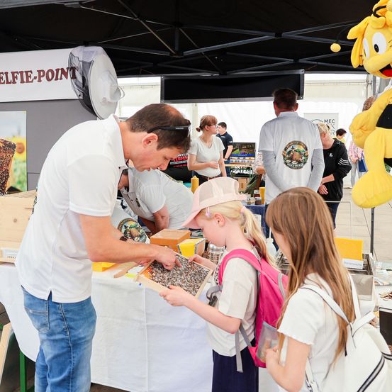 Ein Mann zeigt zwei jungen Mädchen eine Holzkiste mit etwas bei einer Outdoor-Veranstaltung. Dahinter steht ein Tisch mit verschiedenen Gegenständen und einem Maskottchen.
