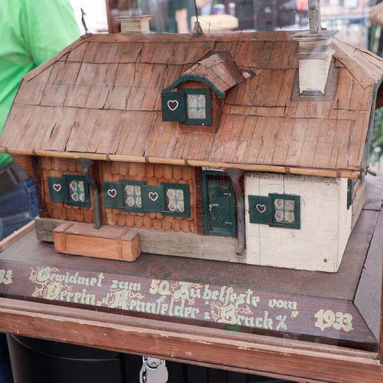 Ein Miniaturhaus aus Holz mit einem Ziegeldach, Holzläden und roten Herzfenstern ist auf einem Holzständer ausgestellt. Das Haus hat einen Schornstein auf der rechten Seite und ist von grünem Gras umgeben. Der Text auf dem Haus lautet: '50. Jahrestag der Ziegel 1933.'
