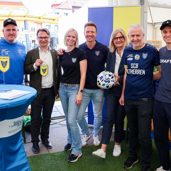 Sieben Personen stehen auf einem grünen Teppich und lächeln für ein Foto. Sie tragen Freizeitkleidung und Turnschuhe. Die Frau in der Mitte hält einen Fußball.