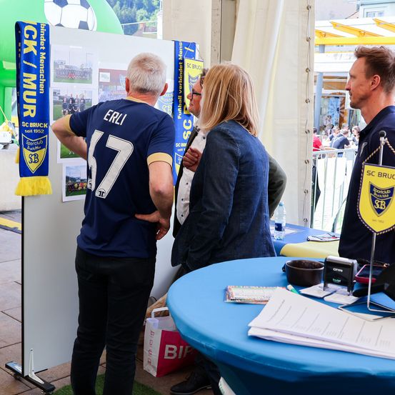 Drei Menschen stehen an einem Tisch, zwei von ihnen tragen Sporttrikots. Der Mann auf der linken Seite trägt ein blaues Trikot mit der Nummer 17 und dem Wort Erli darauf. Die Frau auf der rechten Seite hat eine blaue Jacke. Dahinter befindet sich ein Banner mit der Aufschrift SC BRUCK.