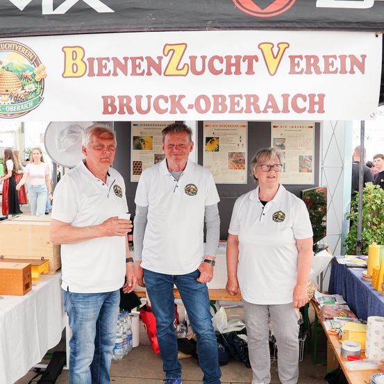 Drei Personen in weißen Polo-Shirts mit Logos stehen vor einem Zelt mit der Aufschrift Bienenzucht Verein Bruck-Oberaich, mit Gegenständen auf einem Tisch und einer Menschenmenge im Hintergrund.