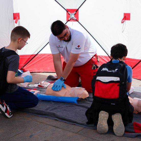 Ein Mann in einem weißen T-Shirt und roten Hosen unterrichtet zwei Jungen, wie man einem Mannequin unter einem Zelt CPR beibringt.