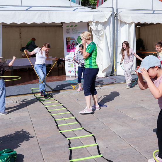 Eine Gruppe von Kindern und einem Erwachsenen macht draußen Übungen. Sie benutzen eine grüne Leiter und Hula-Hoops. Ein Kind klettert die Leiter hoch.